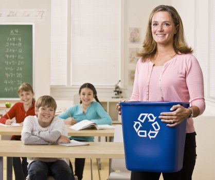 A woman in a pink cardigan holding a blue waste bin with a recycling symbol on the front, standing in a classroom with three children seated at desks, smiling and facing forward. The background shows educational posters, a chalkboard with mathematical equations, and a bright, well-lit environment typical of a school in Chiswick. The scene reflects a focus on environmental awareness and waste management, with the bin positioned prominently in the foreground, suggesting a presentation or activity related to recycling and rubbish reduction to students, in a setting that emphasizes educational and community engagement in London.