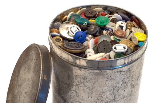 A cylindrical metal container filled with a diverse collection of buttons in various sizes, shapes, and colours, including black, white, blue, red, green, and beige, with some buttons having four holes and others having two. The container, which appears to be made of stainless steel or similar material, has a slightly weathered surface and is positioned outdoors on a paved surface, with a matching lid resting against its side. The background shows a blurred outdoor environment, possibly part of a street or courtyard, indicating a setting related to rubbish or waste collection. The brightness of the image suggests daylight conditions, emphasizing the contrasting textures of the shiny metallic container and the matte surfaces of the buttons. This detailed visual capture of mixed waste items aligns with services offered by rubbish removal companies in the Chiswick area, such as Office Clearance Chiswick, ensuring accurate representation of waste materials encountered during clearance operations.