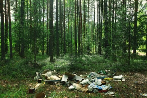 A collection of assorted waste materials including broken furniture, cardboard boxes, plastic bags, and miscellaneous debris is scattered across the forest floor in a wooded area, with tall, slender trees and lush green foliage in the background. The waste appears to be abandoned and untidily piled, contrasting with the natural environment. The scene is illuminated by natural daylight filtering through the canopy, highlighting the disarray of rubbish in the midst of the greenery. This visual depicts improper waste disposal in a woodland setting, relevant to the services provided by Office Clearance Chiswick for rubbish removal and site clearance in the Chiswick area nearby London postal code W4, emphasizing the importance of professional rubbish clearance in preserving natural spaces.