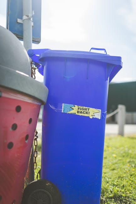 A black wheelie bin labeled with 'St. John's' positioned on a city street at night, filled with various discarded items including cardboard boxes, plastic wrappers, and paper, some of which are partia