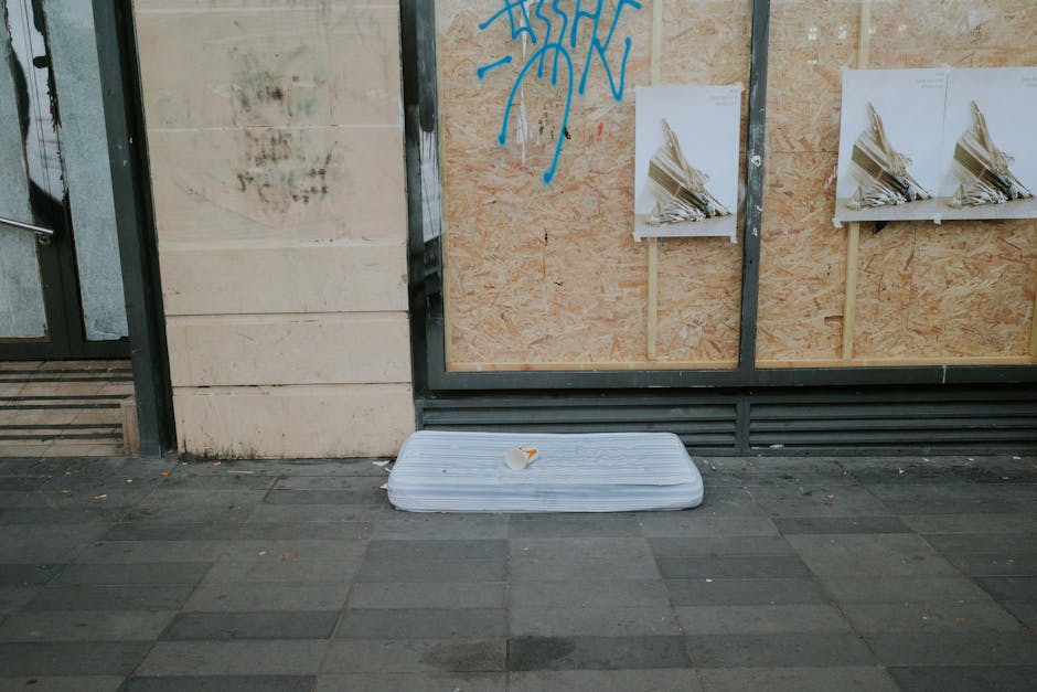 A discarded white foam mattress with a slightly bent edge lies on a dark grey tiled pavement in front of a boarded-up storefront. The mattress appears to be used and has some dirt and stains on its su